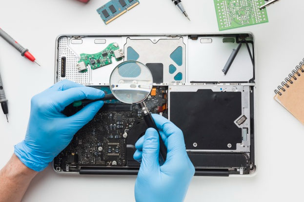 Técnico de luvas inspecionando a placa interna de um notebook aberto, com memória e ferramentas sobre a bancada